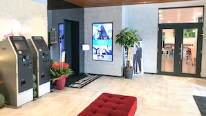A hotel lobby with a reception desk, a red cushioned bench, a digital information kiosk, a large poster, and a potted plant. - Hotel Livemax Fukushima Koriyama Ekimae (Koriyama)
