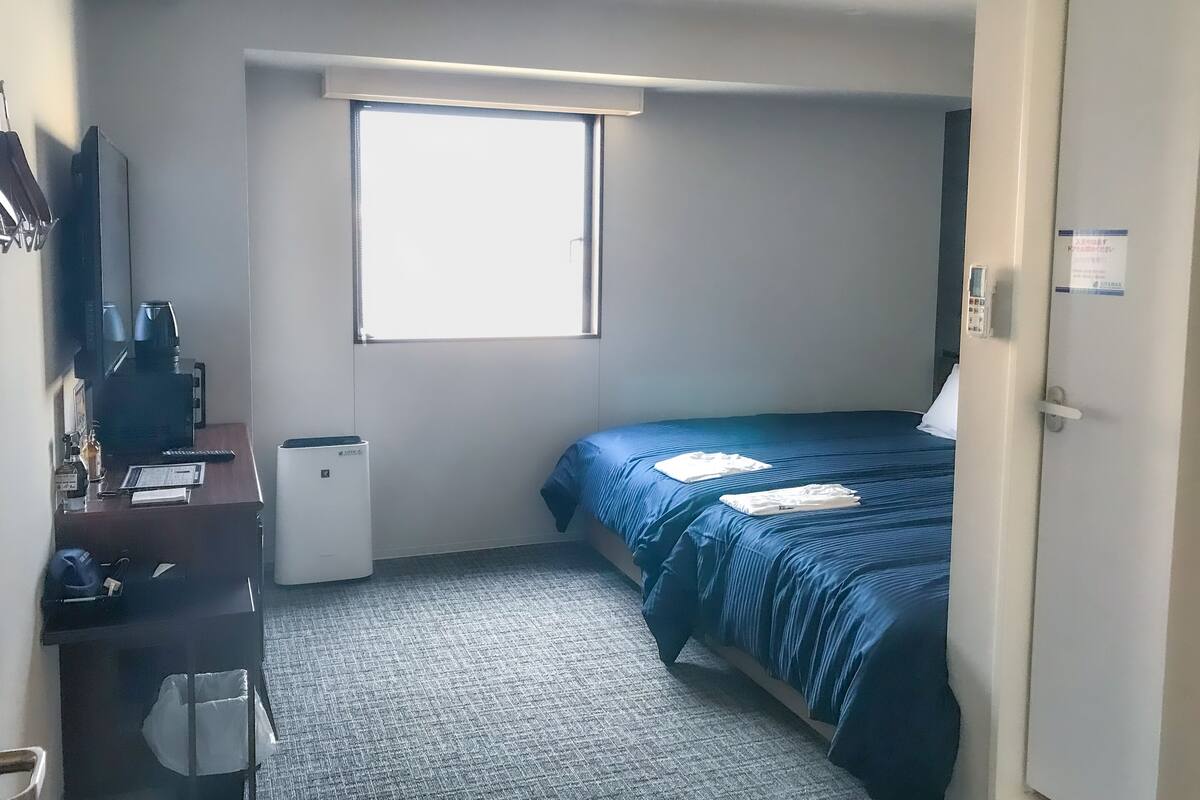 A compact hotel room with a single bed, a desk with a computer, a small heater, and a window providing natural light.