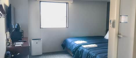A compact hotel room with a single bed, a desk with a computer, a small heater, and a window providing natural light.
