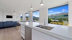 Une cuisine moderne dotée d’un grand îlot central, de meubles blancs et d’un lave-vaisselle. On y trouve également des luminaires suspendus, un téléviseur à écran plat et de grandes fenêtres offrant une vue panoramique sur les montagnes et un village.