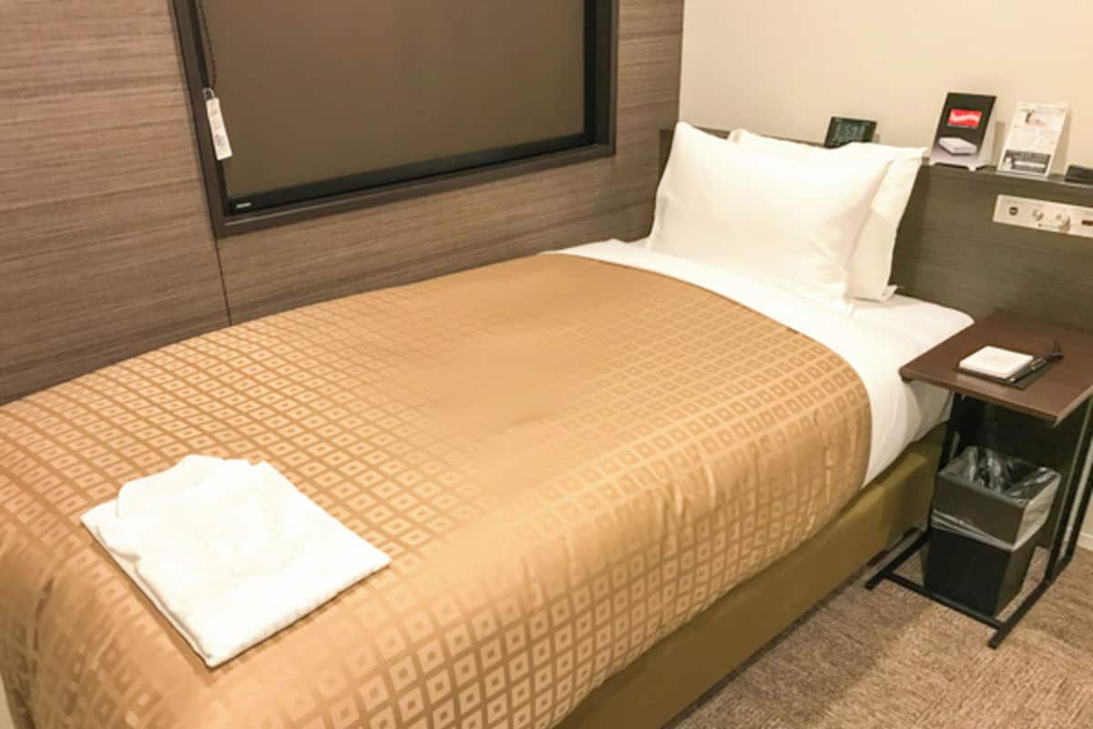 A hotel room with a bed, a nightstand, a trash bin, and a wall-mounted control panel.