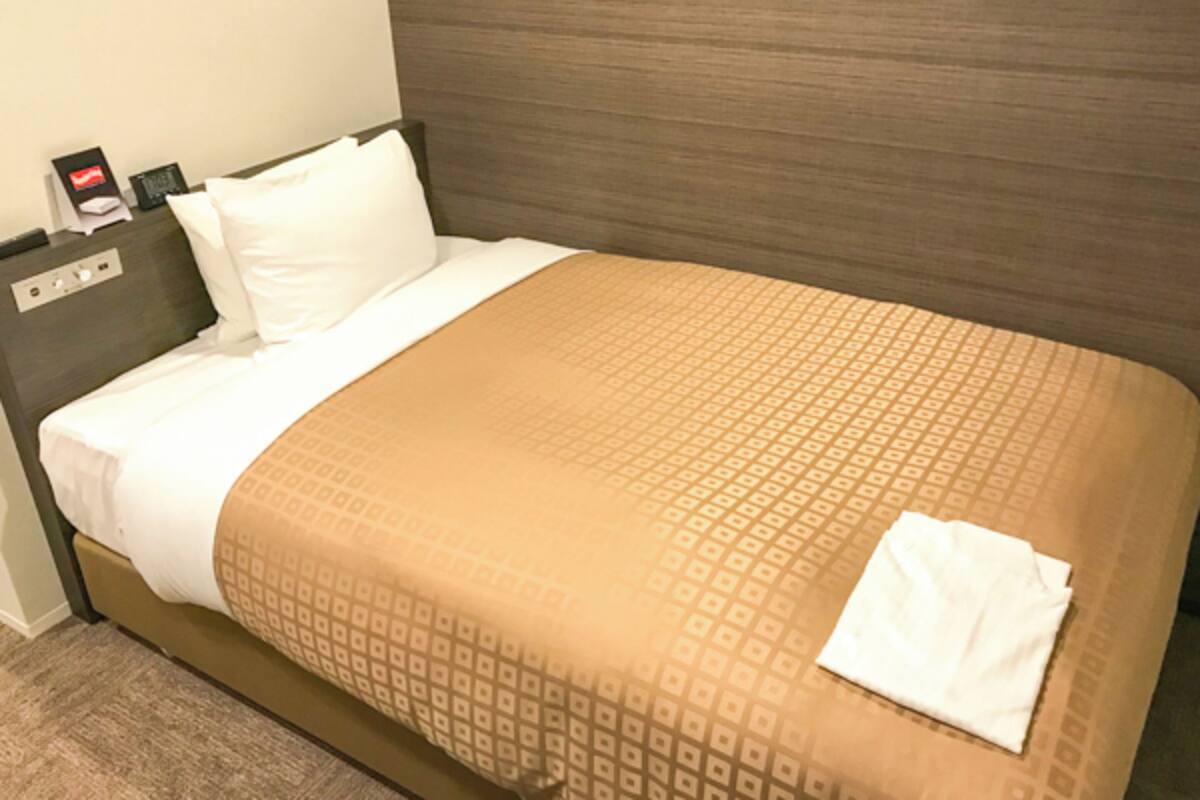 A hotel room with a bed, a nightstand, and a wall-mounted control panel.