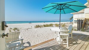 Vista a la playa desde una terraza de madera con un paraguas turquesa, sillas blancas y una puerta con cerradura.