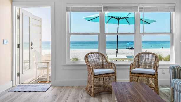 Una habitación con un gran ventanal con vistas a la playa, sillas de mimbre, una mesa de madera y un sofá a rayas.