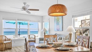 Comedor con mesa de madera dispuesta para cuatro, rodeado de sillas de mimbre, con vista al océano a través de amplios ventanales.