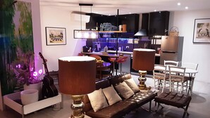 A modern living room with a sofa, two lamps, a dining table, and chairs. There is a kitchen area in the background with a bar and a hanging light fixture. - Duplex garden terrace in the heart of Juan-les-Pins. 6/8 people. (Antibes)