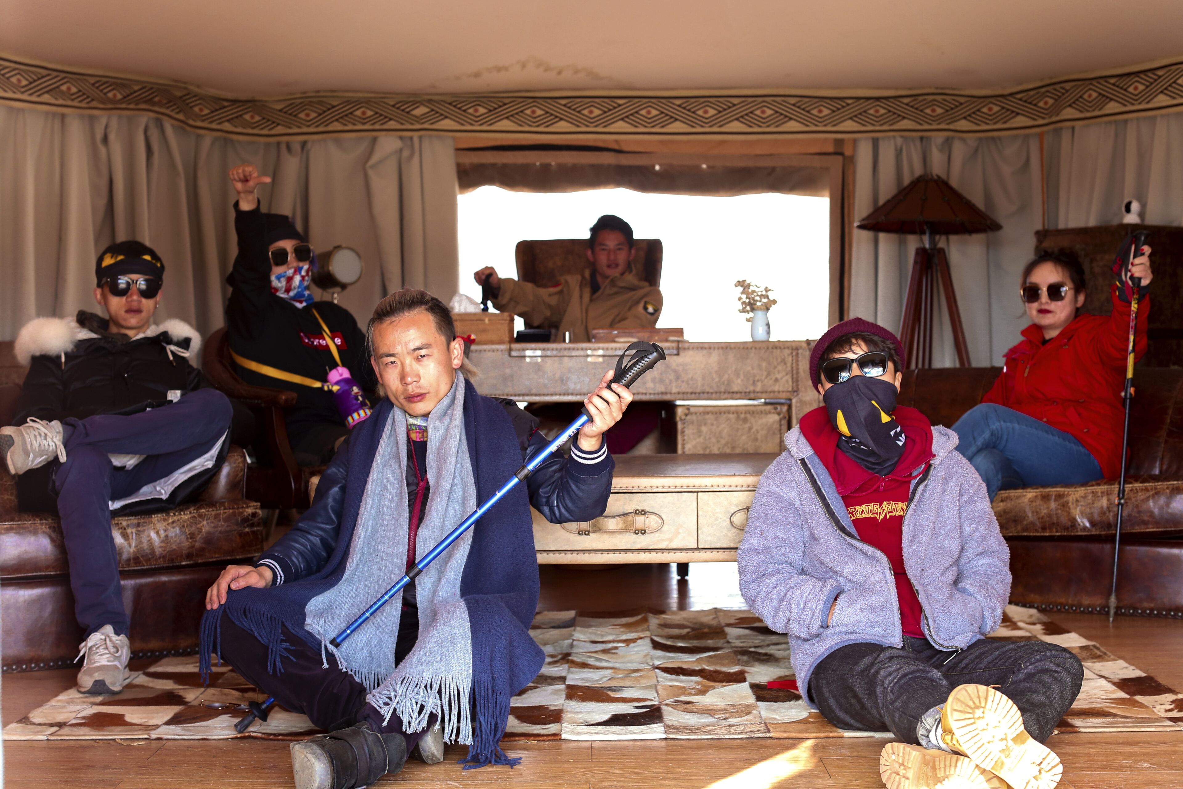 A group of people sitting on a patterned rug in a room with a large window, a lamp, and a wooden cabinet.