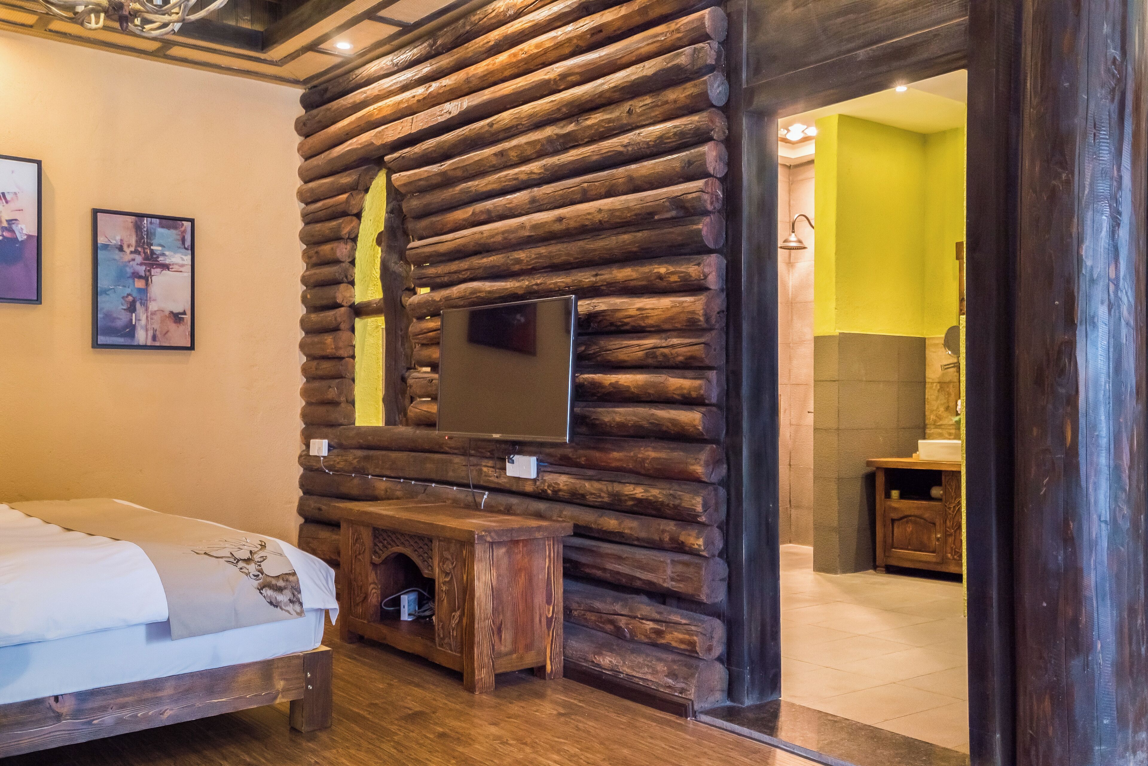 Une pièce aménagée dans le style d’un chalet en rondins, avec un lit, un banc en bois, un téléviseur fixé au mur et une salle de bain visible à travers une porte ouverte.