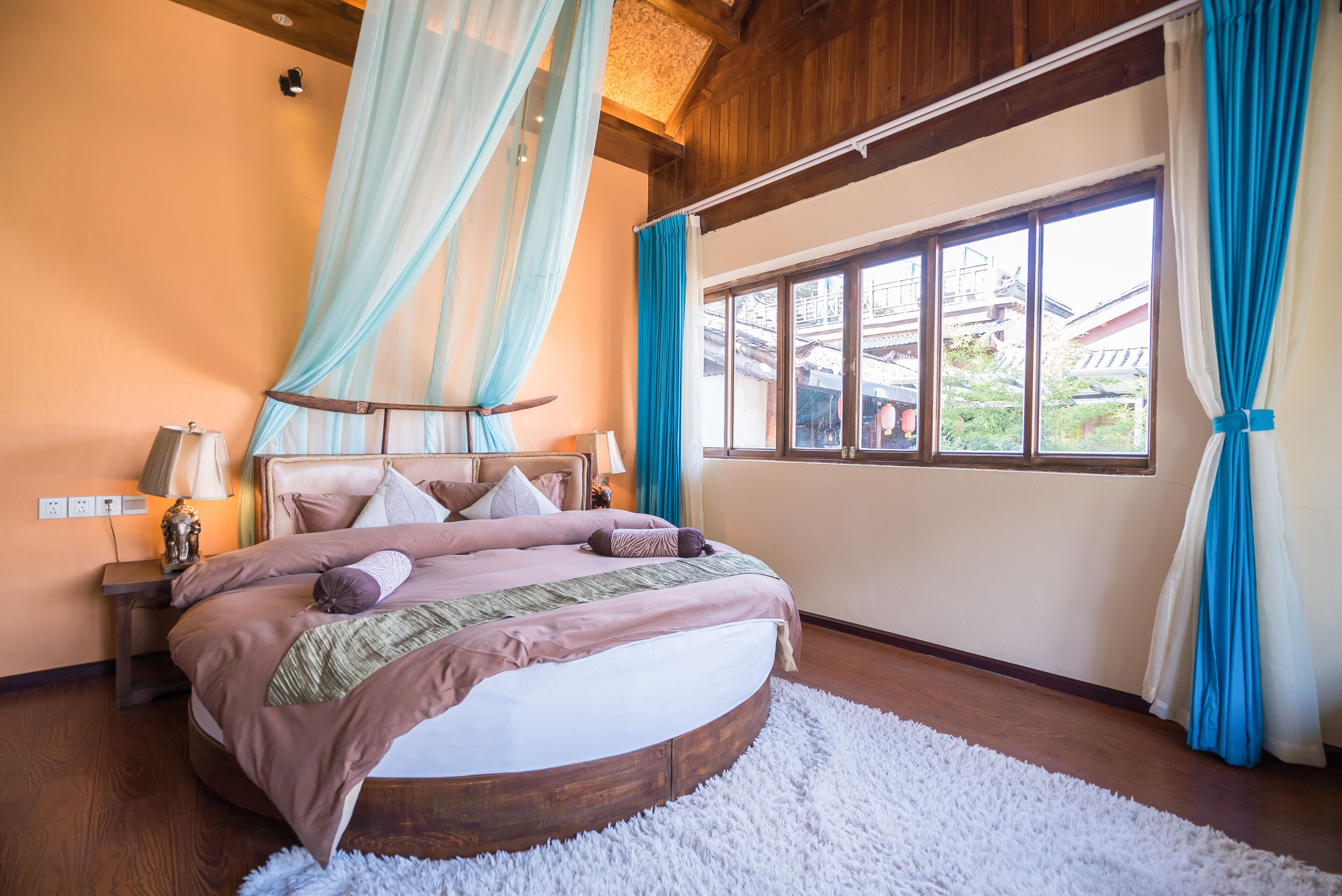 A bedroom with a large bed, a window with curtains, a nightstand with a lamp, and a view of traditional architecture outside.