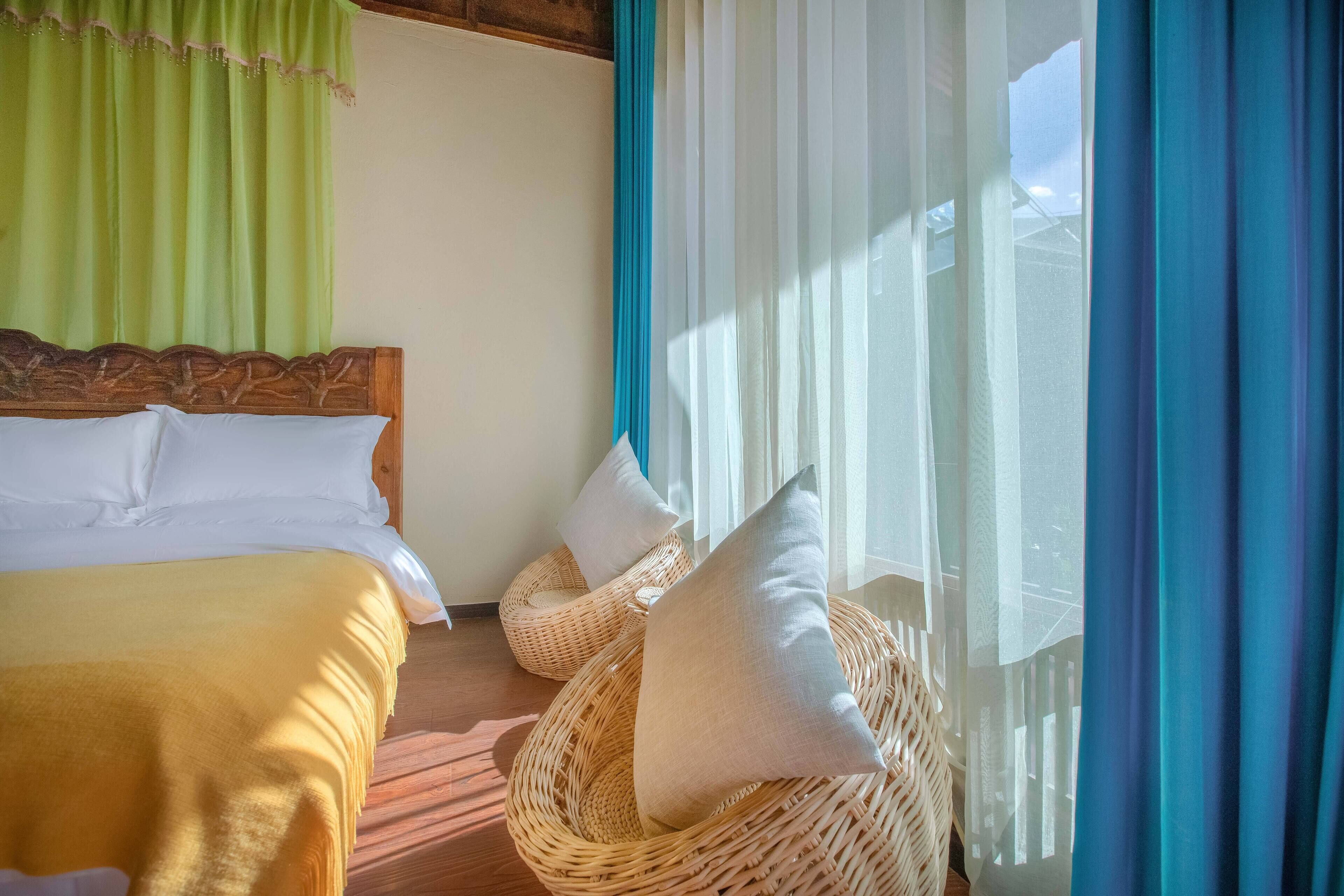A neatly made bed with white linens and a yellow blanket, a wicker chair with a pillow, and sheer curtains allowing natural light.