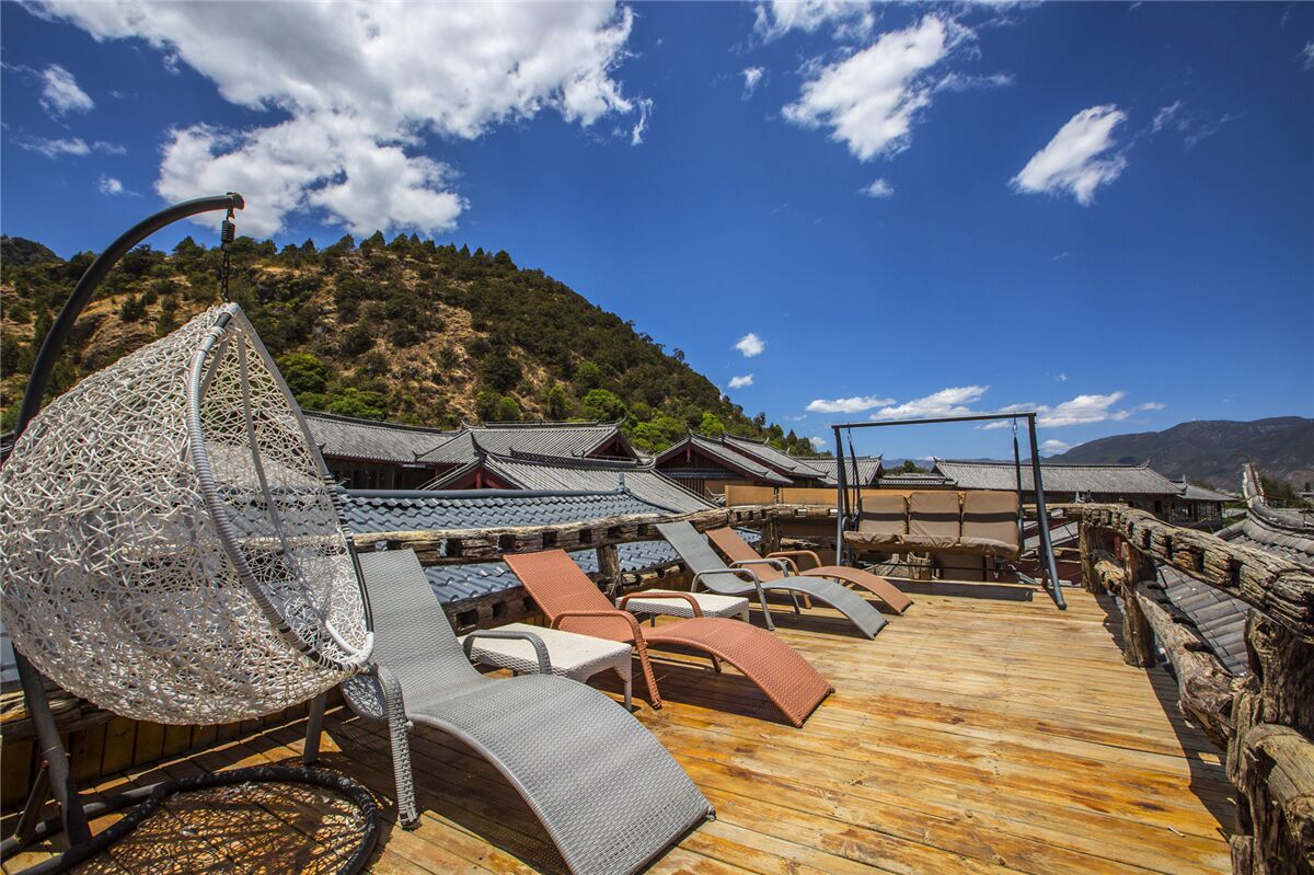 A wooden deck with lounge chairs and a decorative metal chair, overlooking traditional buildings and a mountainous landscape.