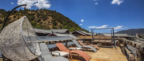 A wooden deck with lounge chairs and a decorative metal chair, overlooking traditional buildings and a mountainous landscape.