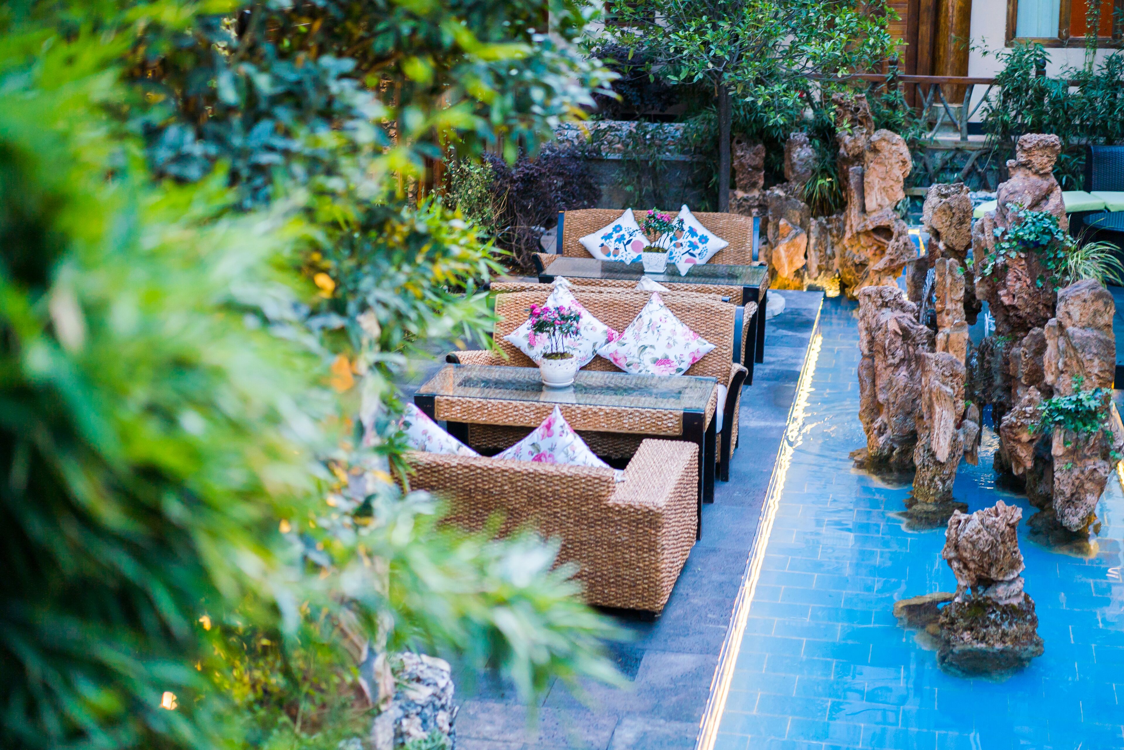 Un espace extérieur paisible, aménagé avec des meubles en osier et des coussins décoratifs, entouré d’une végétation luxuriante et agrémenté d’un jardin de rocaille.