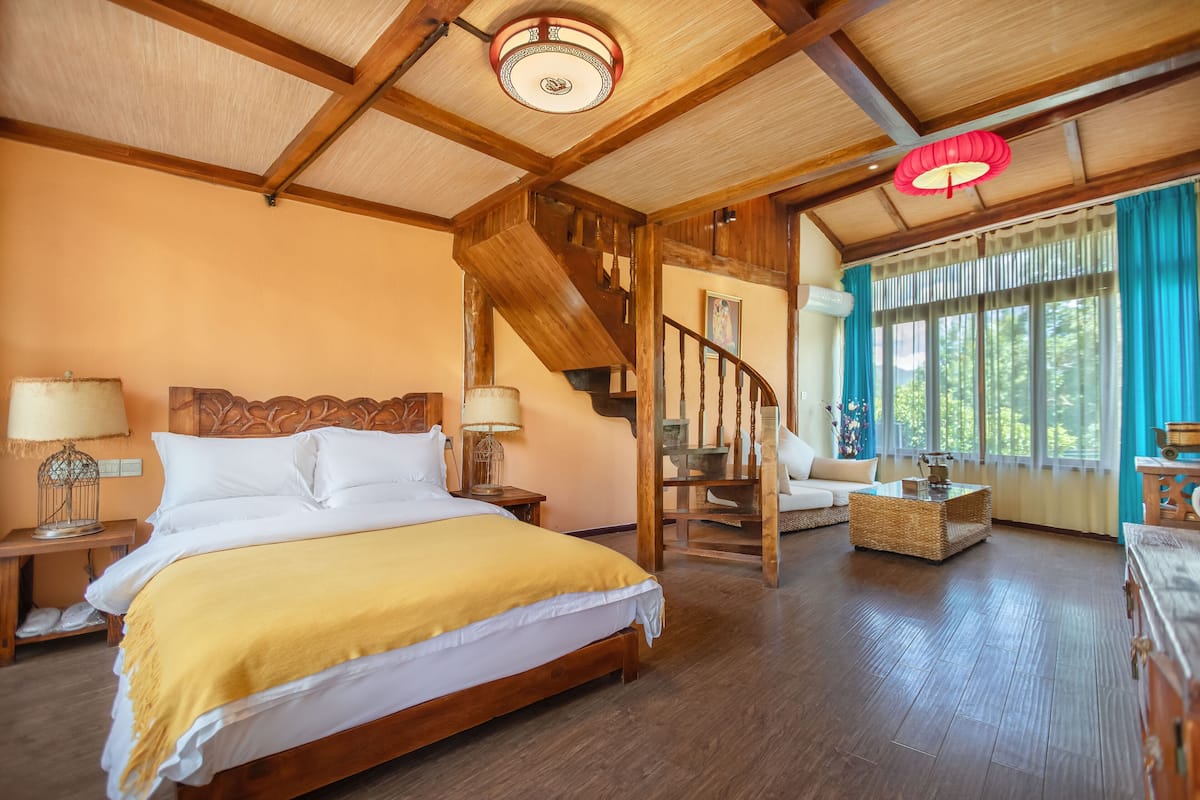 Ein geräumiges Schlafzimmer mit einem großen Bett, Holzbalkendecke, einer Treppe und Blick ins Freie durch ein Fenster.