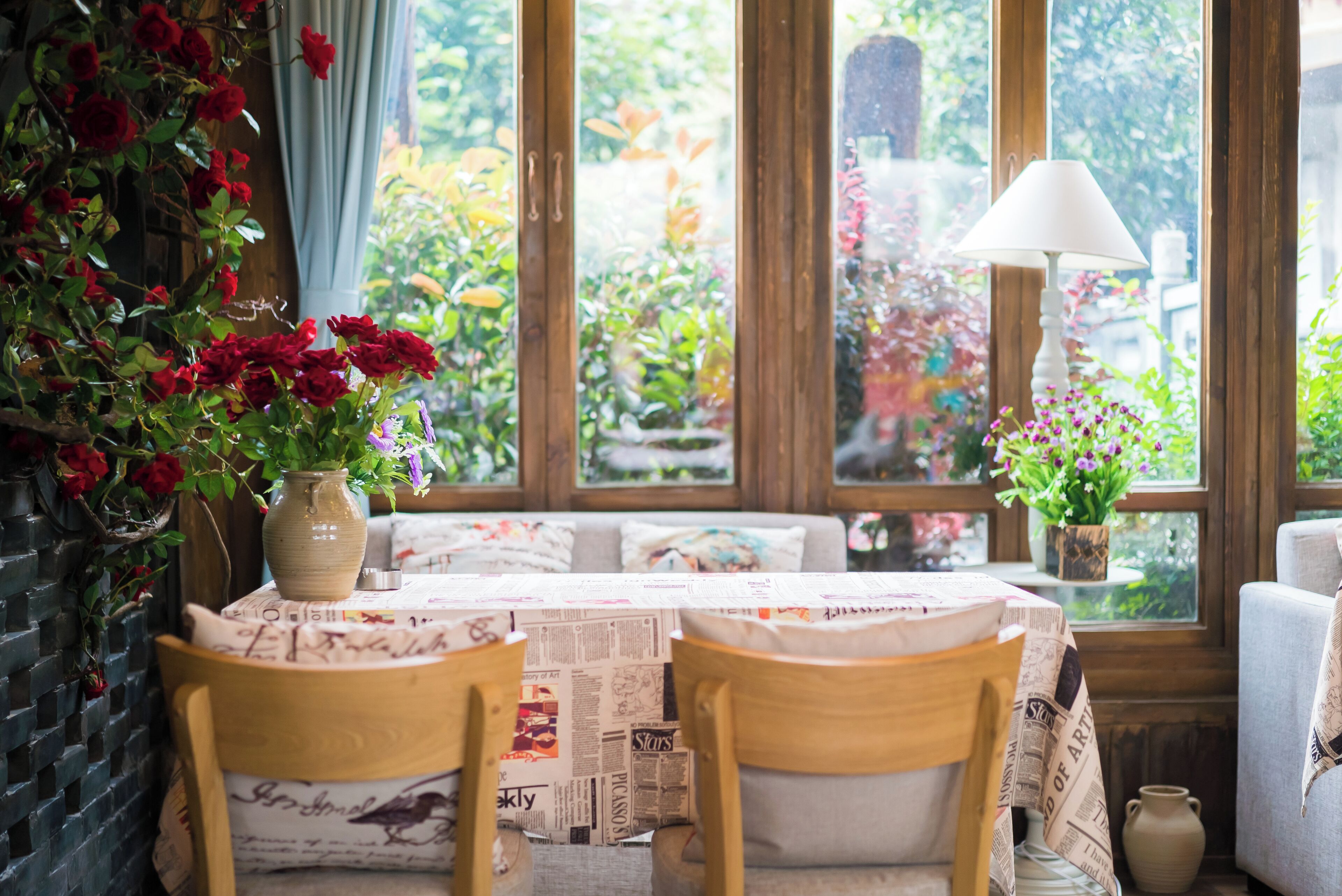 A dining table with newspaper tablecloths, two wooden chairs, a vase with red flowers, and a lamp on a windowsill.