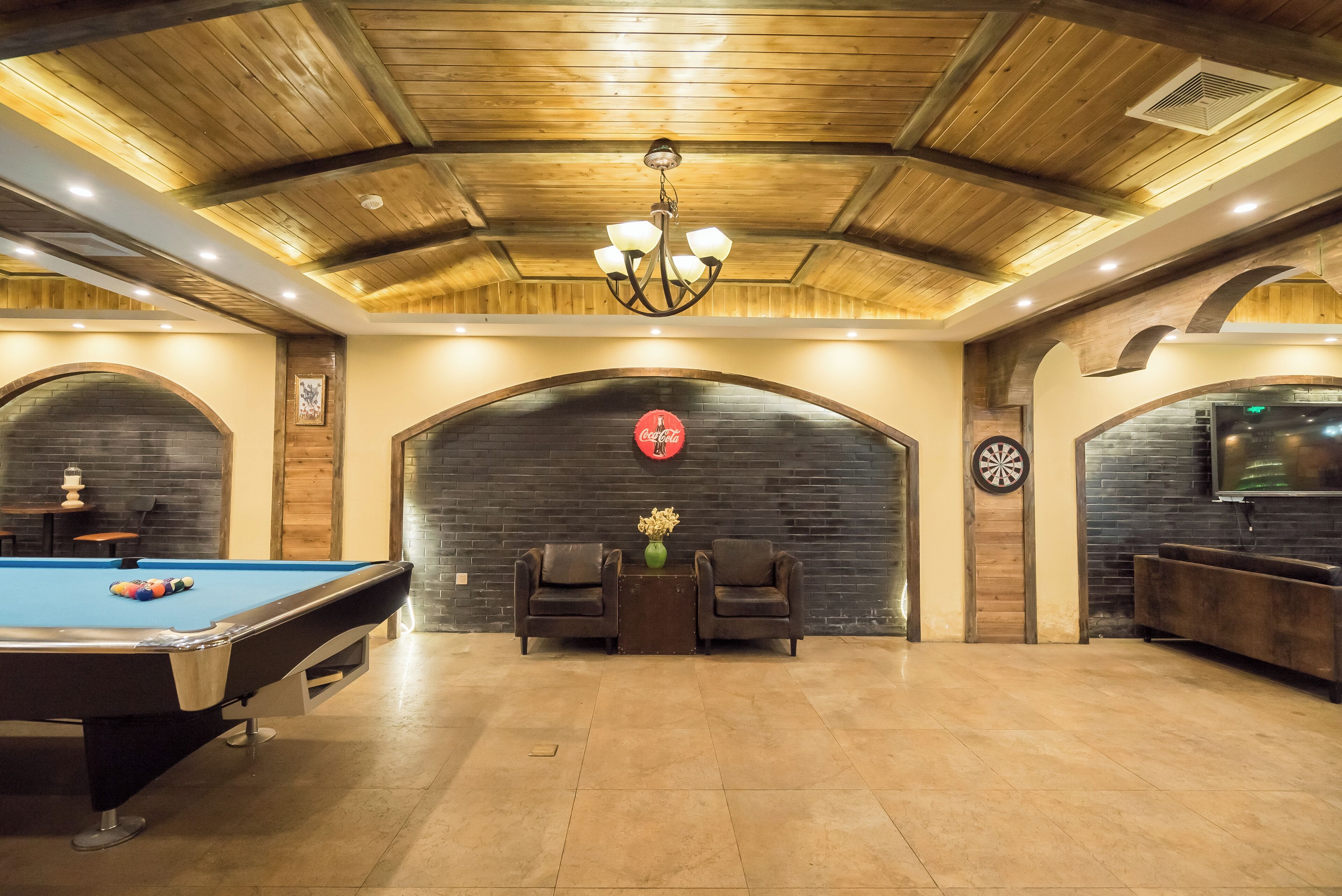 Une table de billard avec un jeu de billard (queue et billes), deux fauteuils en cuir, un jeu de fléchettes et une enseigne Coca-Cola sur un mur en briques.