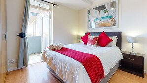 A neatly made bed with white and red bedding, a nightstand with a lamp, and a painting of a beach scene on the wall. - Horizon Bay 405 (Cape Town)