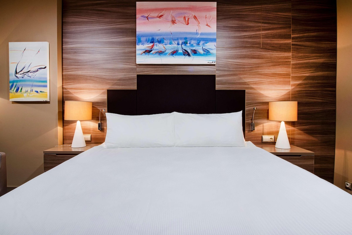 Una habitación de hotel con una cama grande, dos mesitas de noche con lámparas y dos cuadros en la pared.