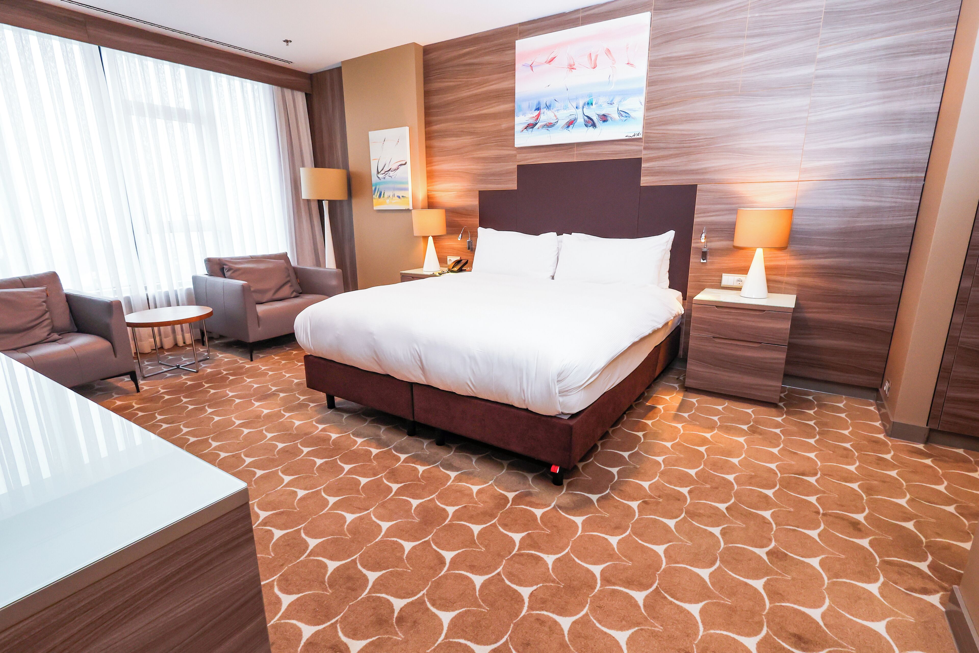 A hotel room with a large bed, a sitting area with a table and chairs, and a view of the outdoors through the window.