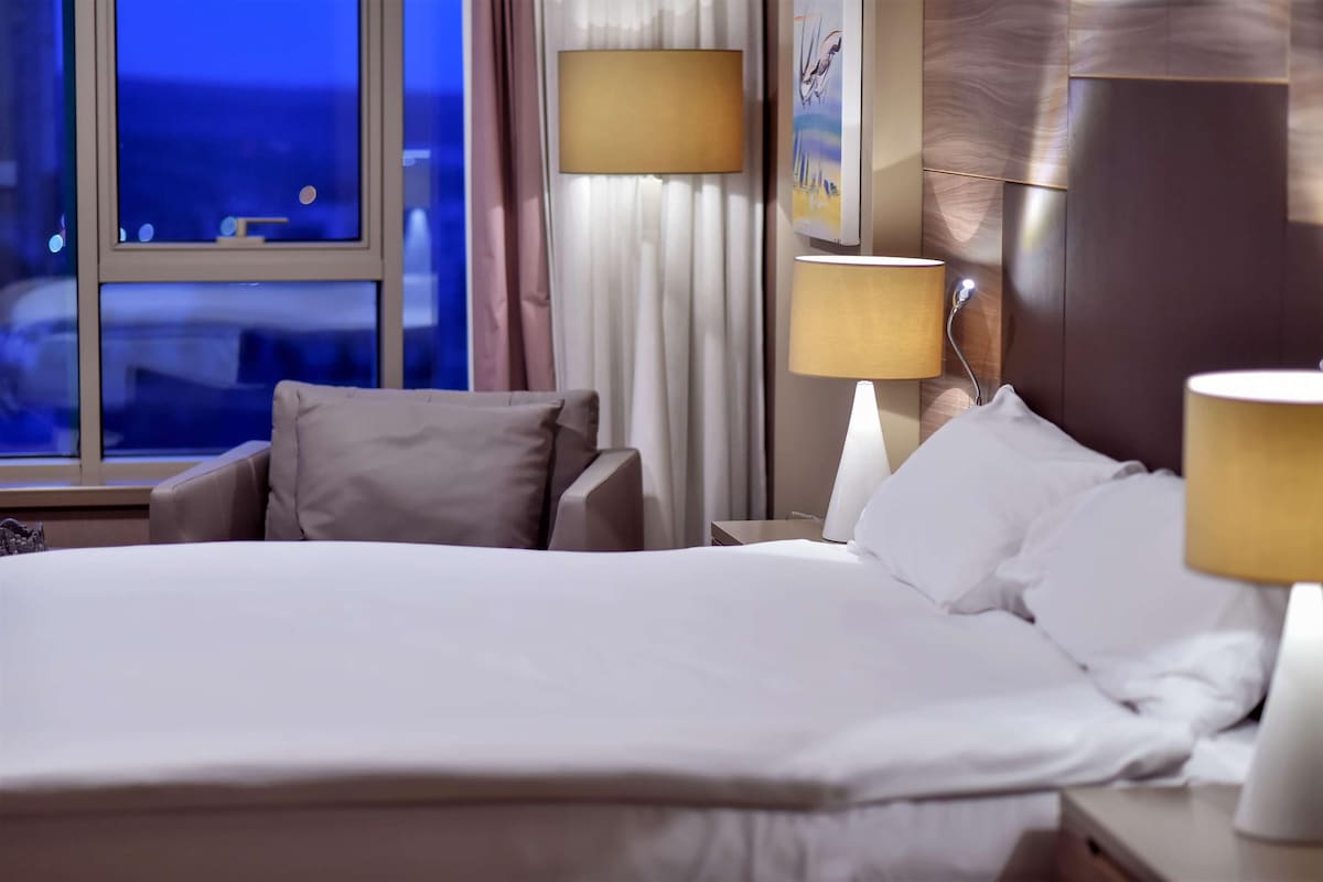 Habitación de hotel con una cama grande, dos lámparas de noche, una mesita de noche y vistas a la ciudad a través de la ventana.