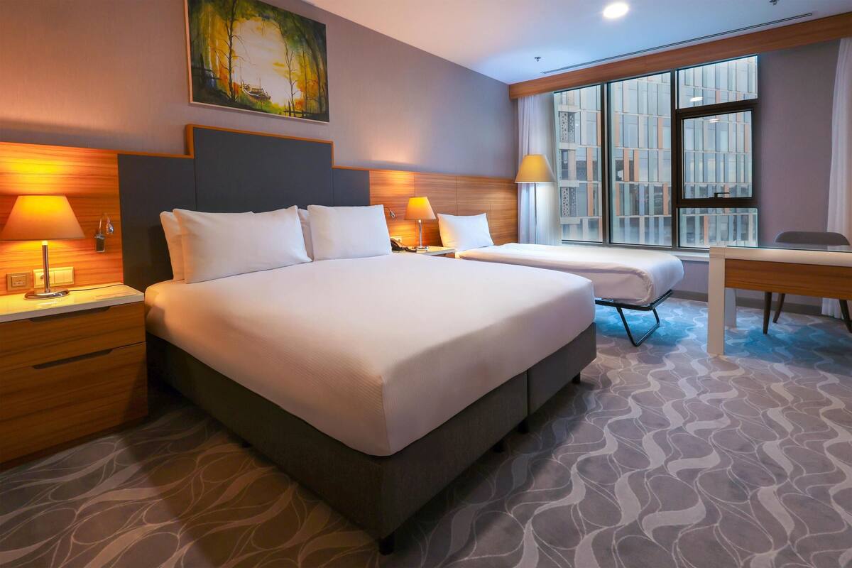 A hotel room with a large bed, two bedside tables with lamps, a desk, and a large window with a city view.