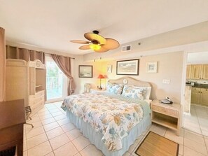 A bedroom with a bed, a nightstand, a lamp, a ceiling fan, and a view of the kitchen.