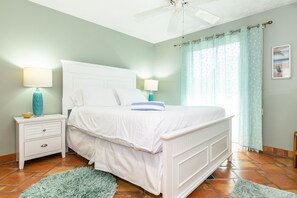 Quarto com cama branca, mesa de cabeceira com abajur, ventilador de teto e janela com cortinas.