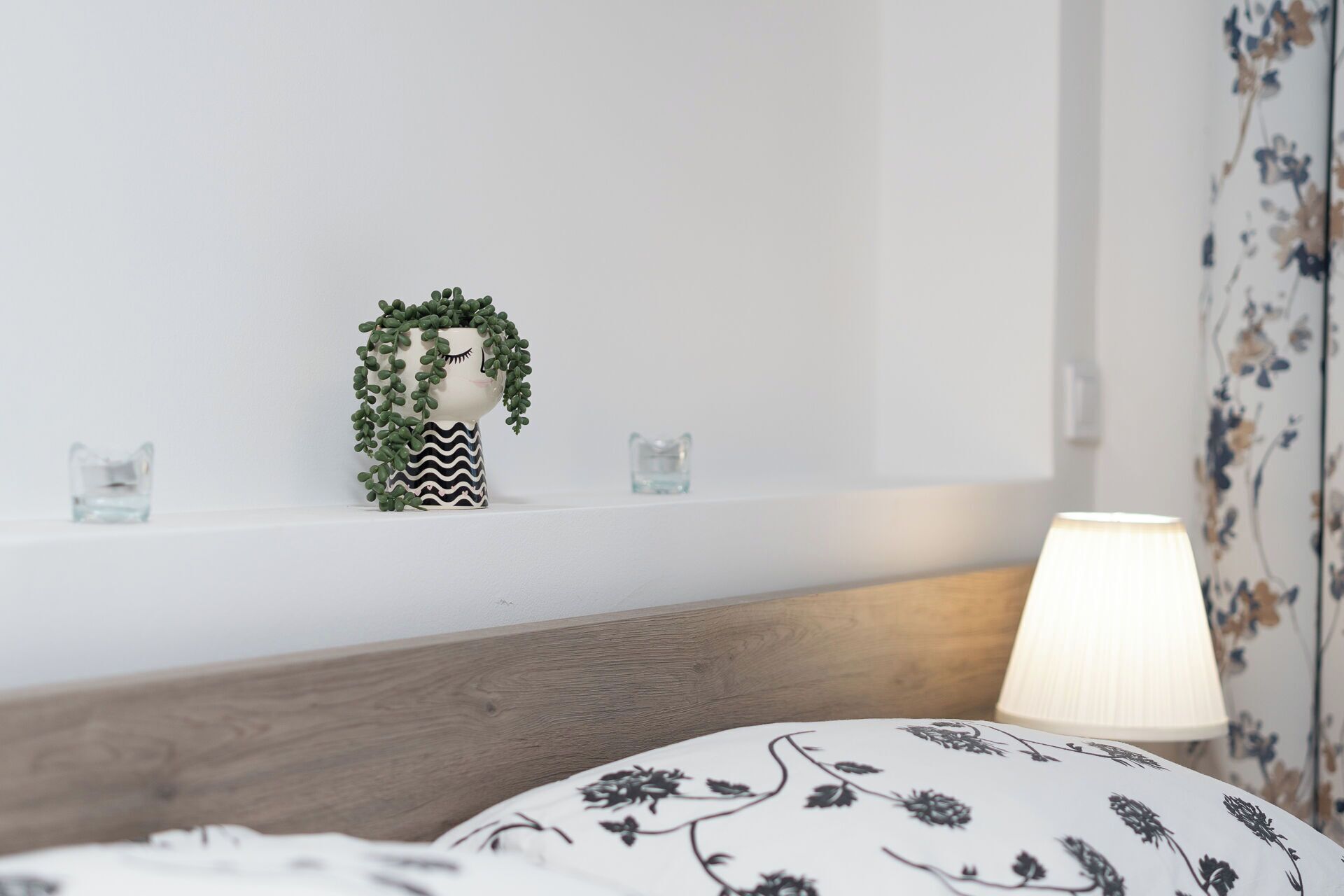 A bedroom with a wooden headboard, a bedside lamp, a floral-patterned pillow, and a shelf with a decorative item and two small glasses.