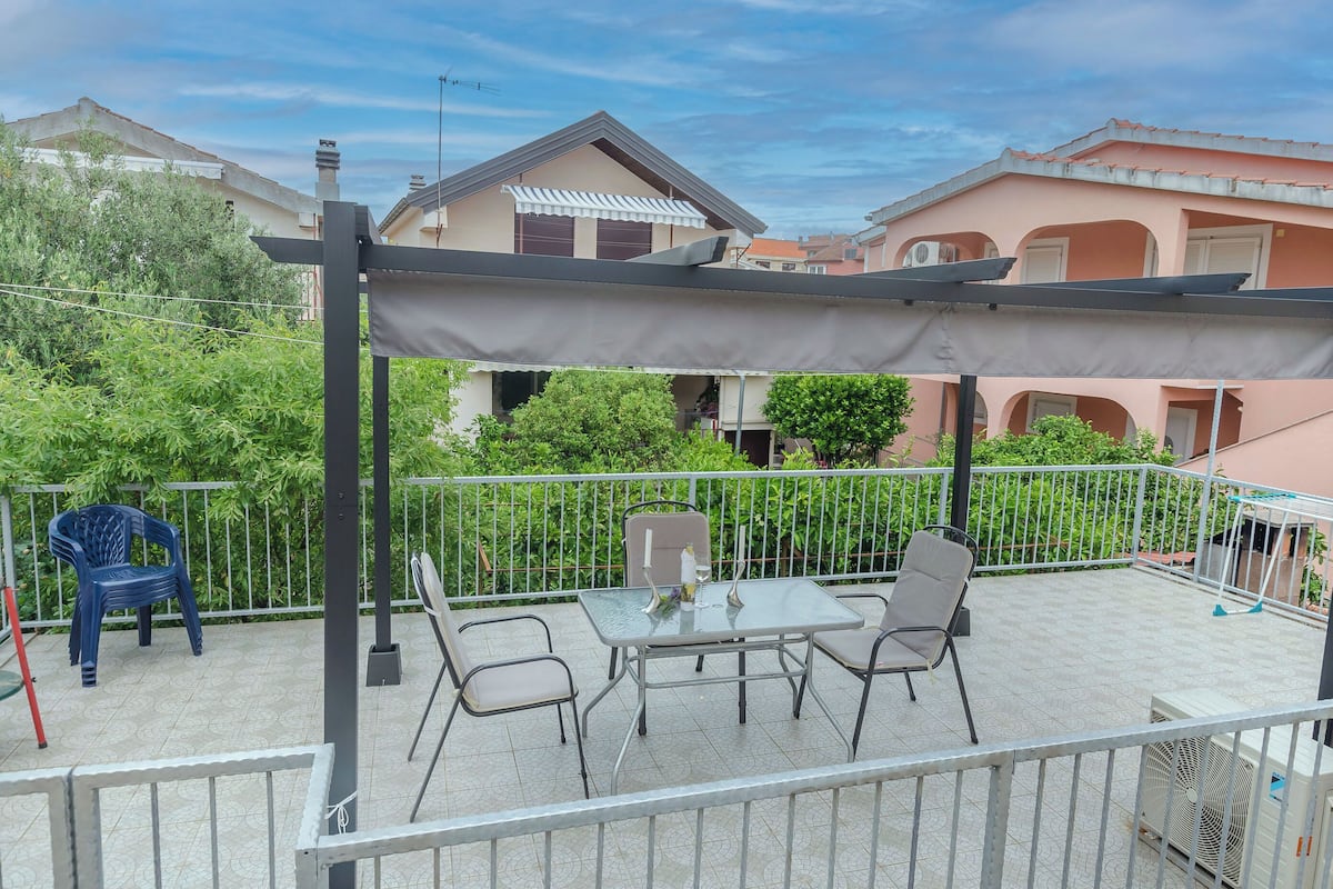 En balkong med ett bord och stolar, en pergola och utsikt över de omkringliggande husen.