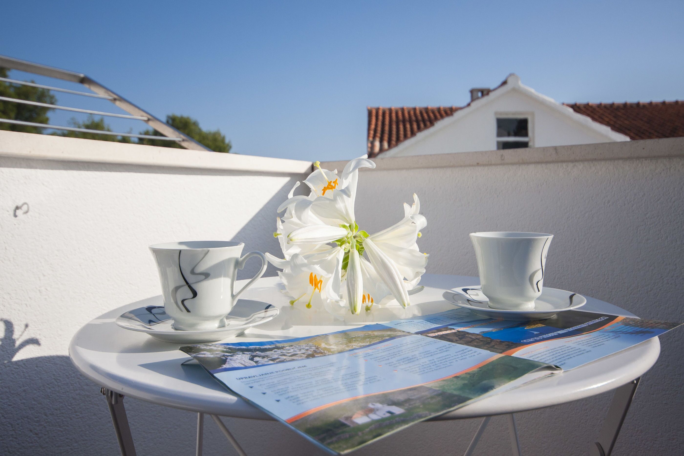 A small round table with a white tablecloth, two white cups, a white teapot, and a white flower arrangement on a balcony.