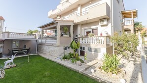 A two-story residential building with a balcony, a small garden area with a lawn and plants, and a patio with outdoor furniture. - Apartments Rajka (Vodice)