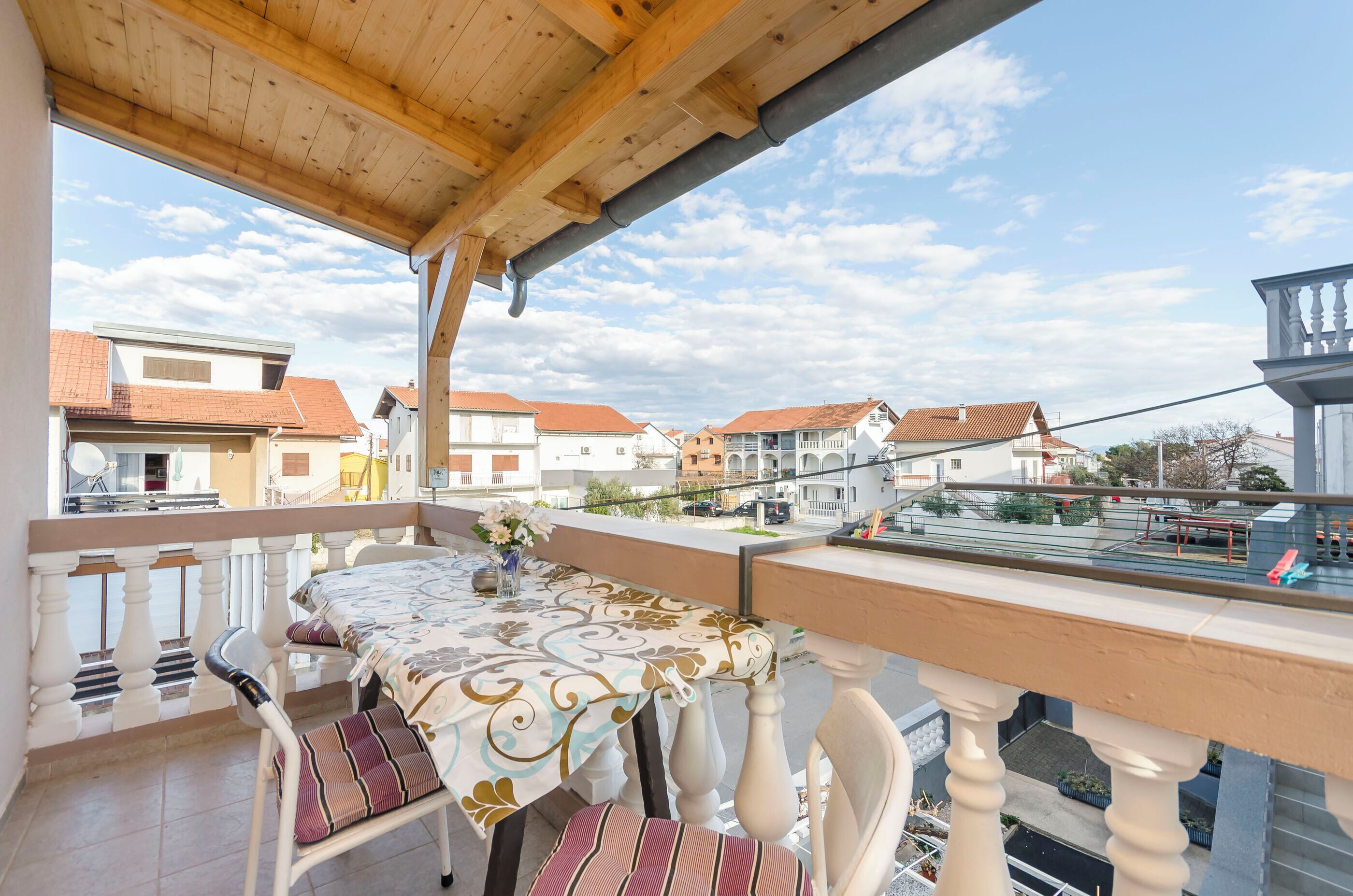 Varanda com mesa e cadeiras, vista para uma área residencial com casas e céu claro.