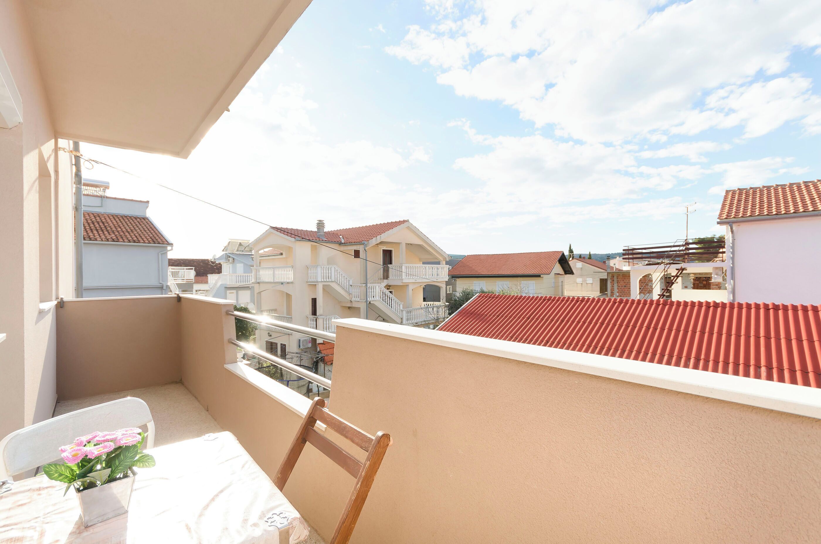 A balcony with a chair and a table, overlooking neighboring buildings with red-tiled roofs.