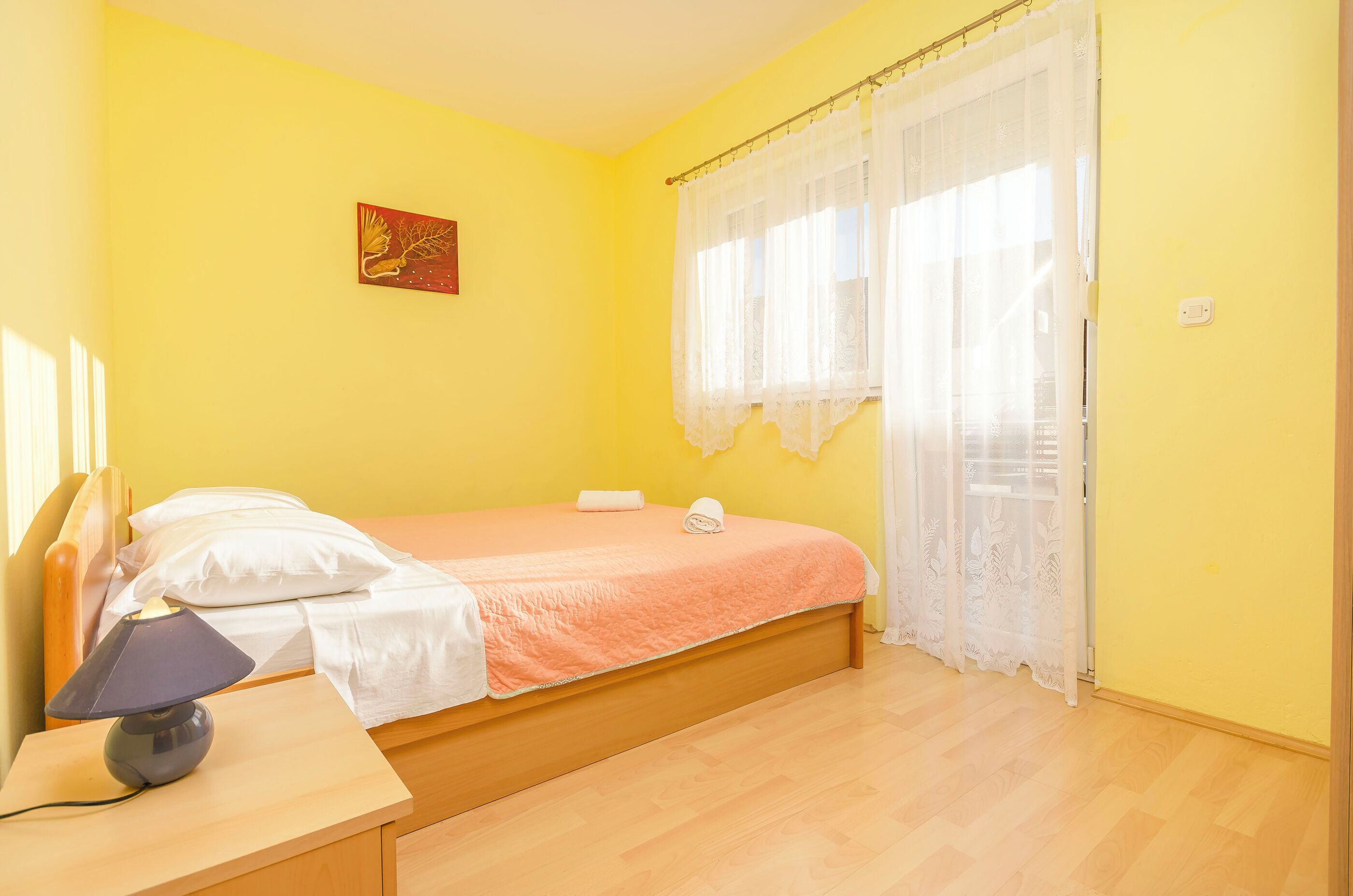 A bedroom with a bed, a nightstand, a lamp, and a window with sheer curtains.