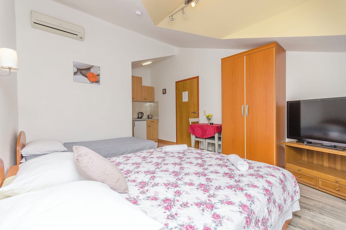 A bedroom with a bed, a TV, a wardrobe, and a kitchenette in the background.
