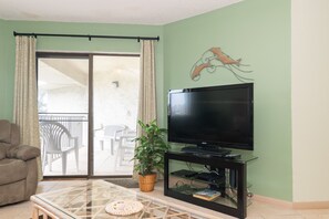 Ein Wohnzimmer mit Flachbildfernseher, Glastisch, Sofa, Topfpflanze und einer Glastür, die zu einem Außenbereich mit Sitzgelegenheiten führt.