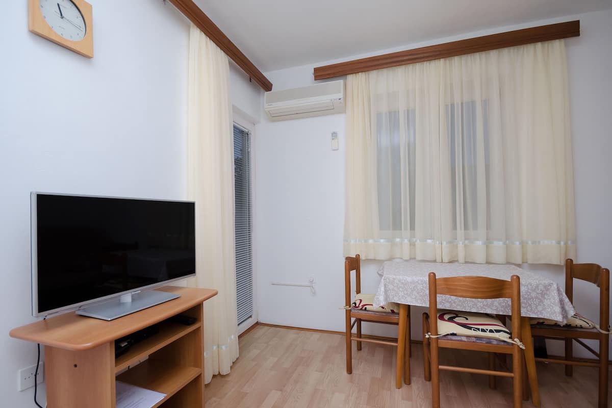 A room with a flat-screen TV on a wooden stand, a table with chairs, a window with sheer curtains, and an air conditioning unit on the wall.