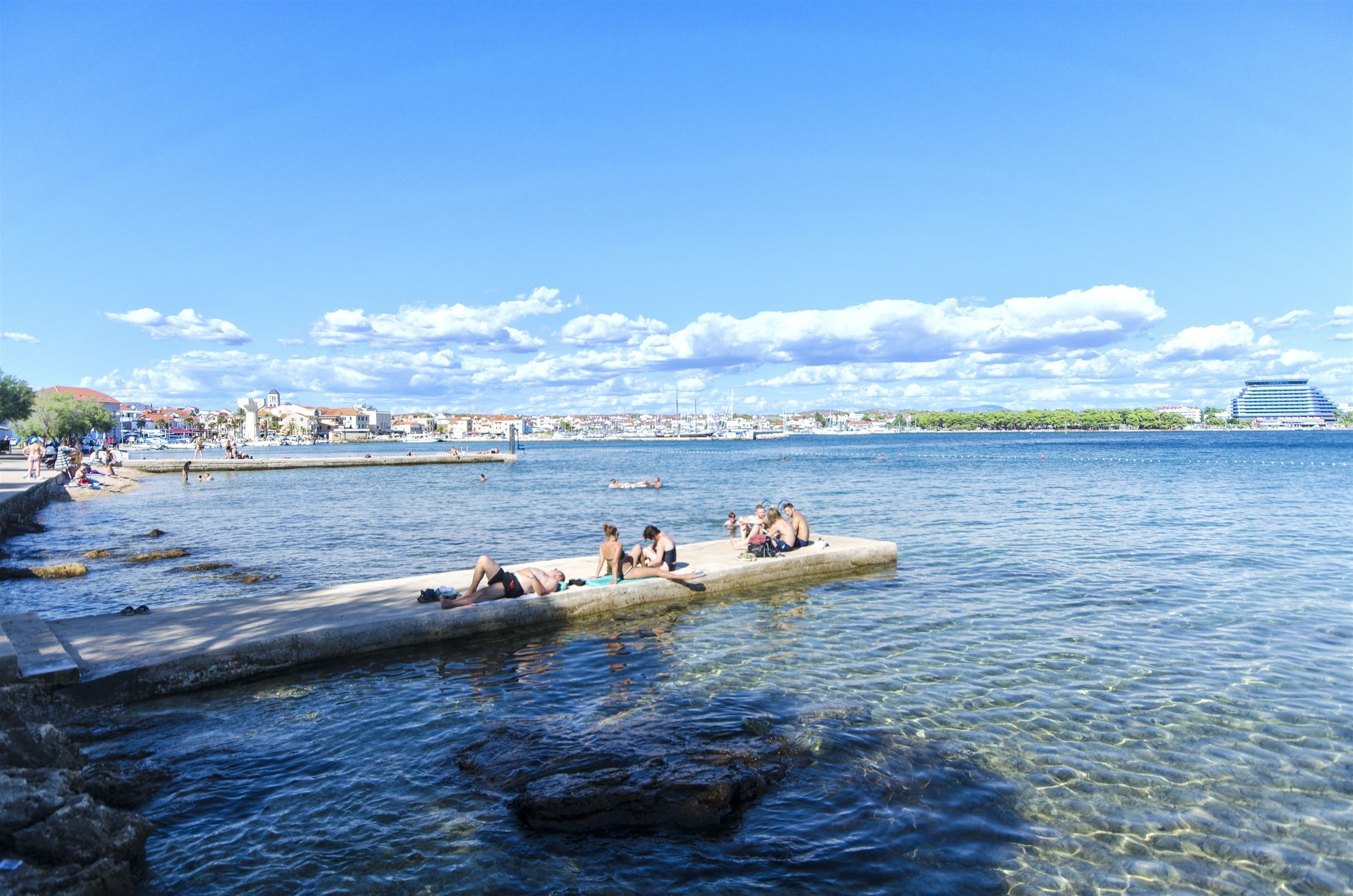A waterfront area with people relaxing on a dock, clear water, and distant buildings.