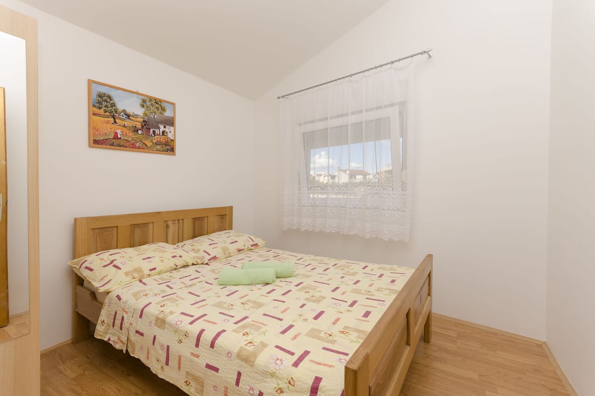 Une chambre avec un lit en bois, une couvre-lit à motifs floraux, un tableau au mur et une fenêtre ornée de rideaux légers.