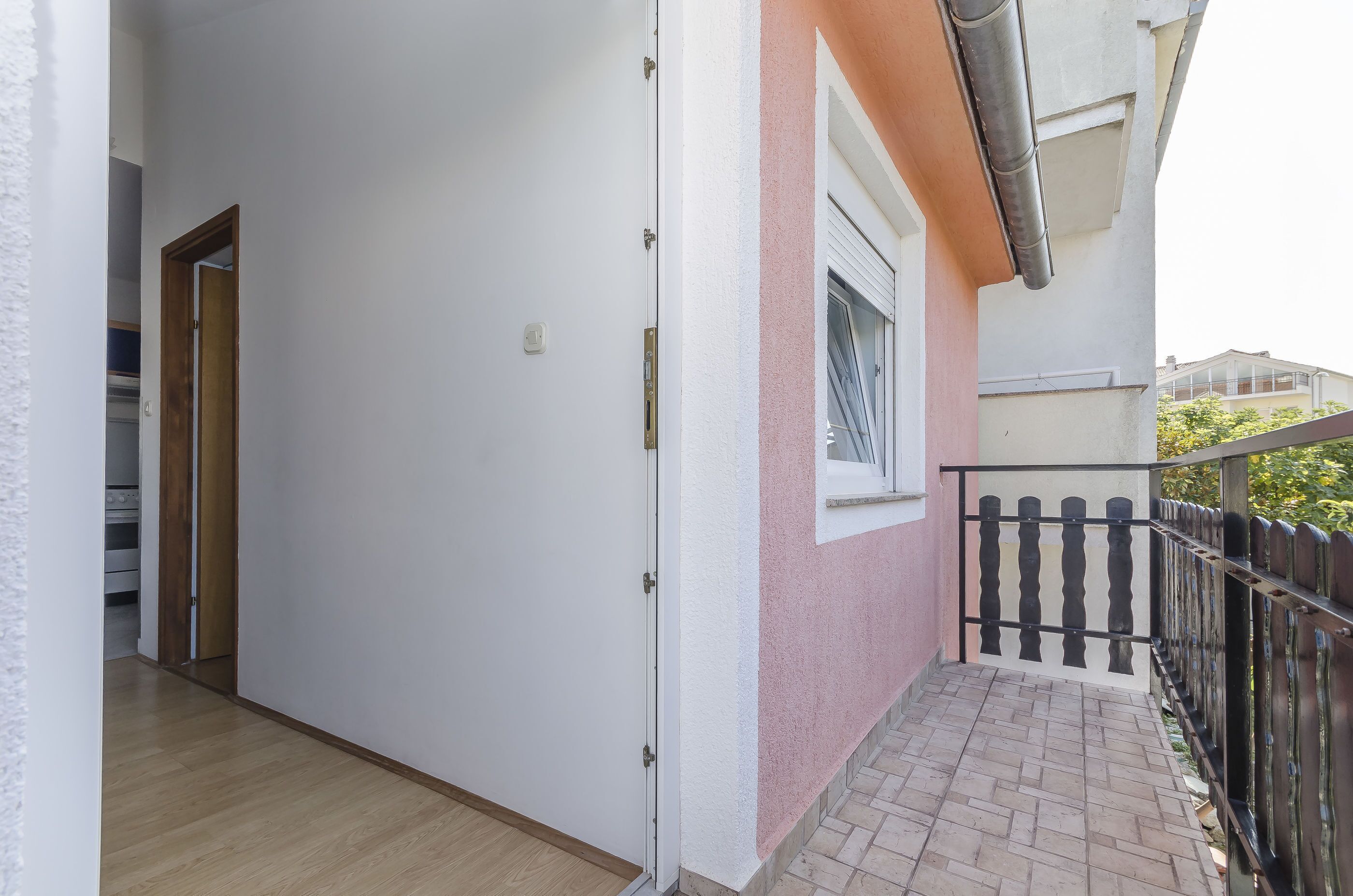 A corridor with a door leading to a room, a balcony with a metal railing, and a window with blinds.