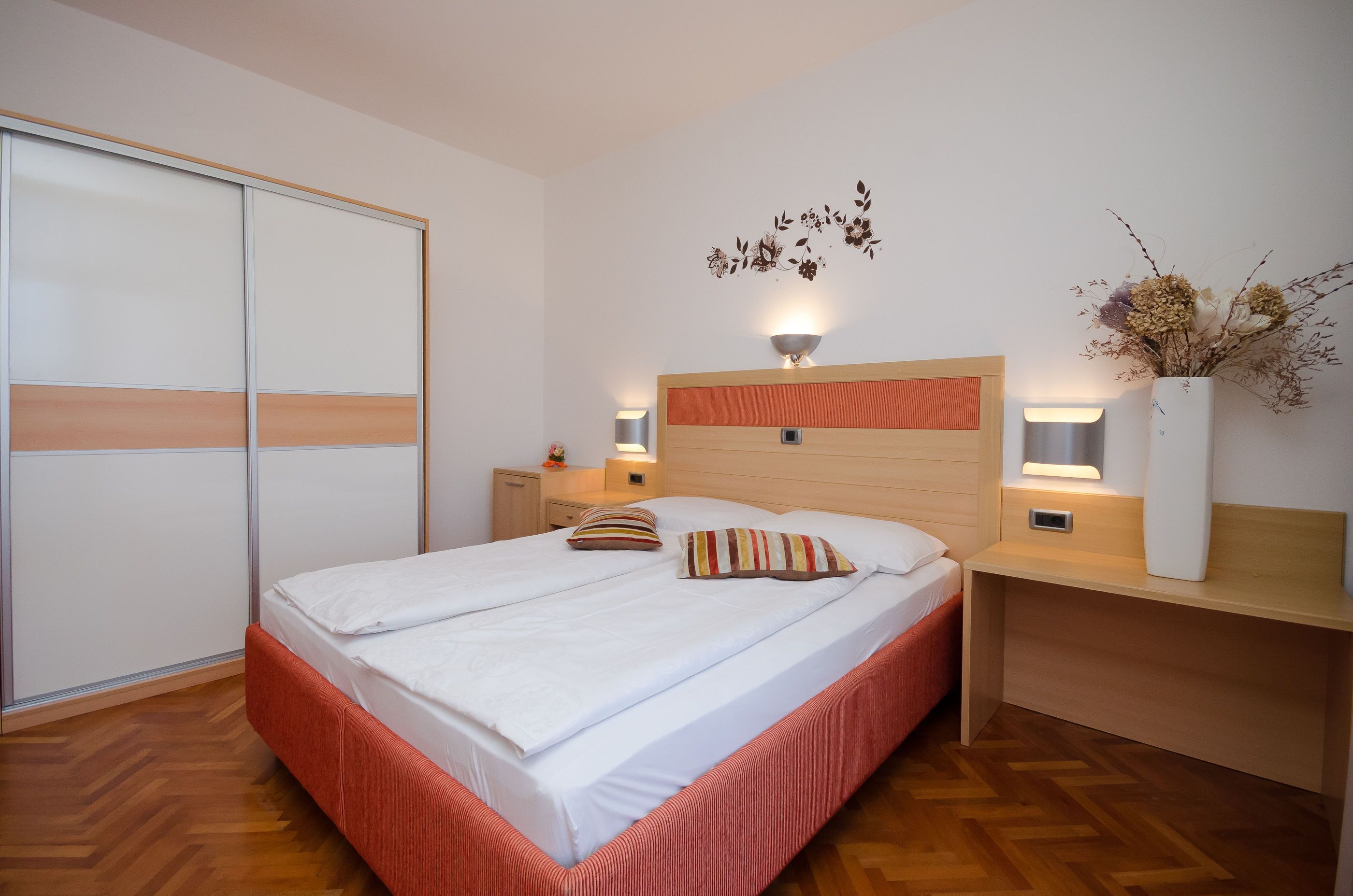 A neatly arranged bedroom with a bed, bedside tables, a nightstand, a wardrobe, and a wall-mounted light fixture.