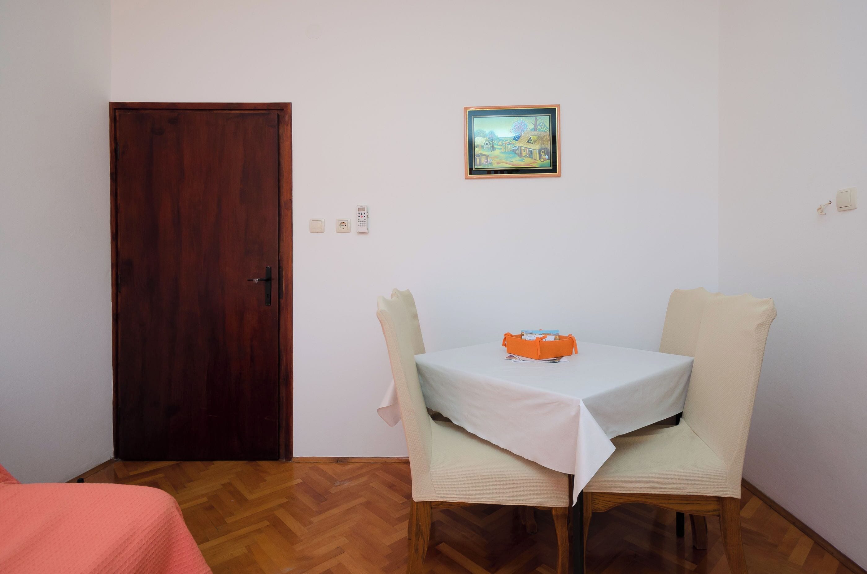 Una habitación con puerta de madera, una mesita con mantel blanco, dos sillas y un cuadro enmarcado en la pared.