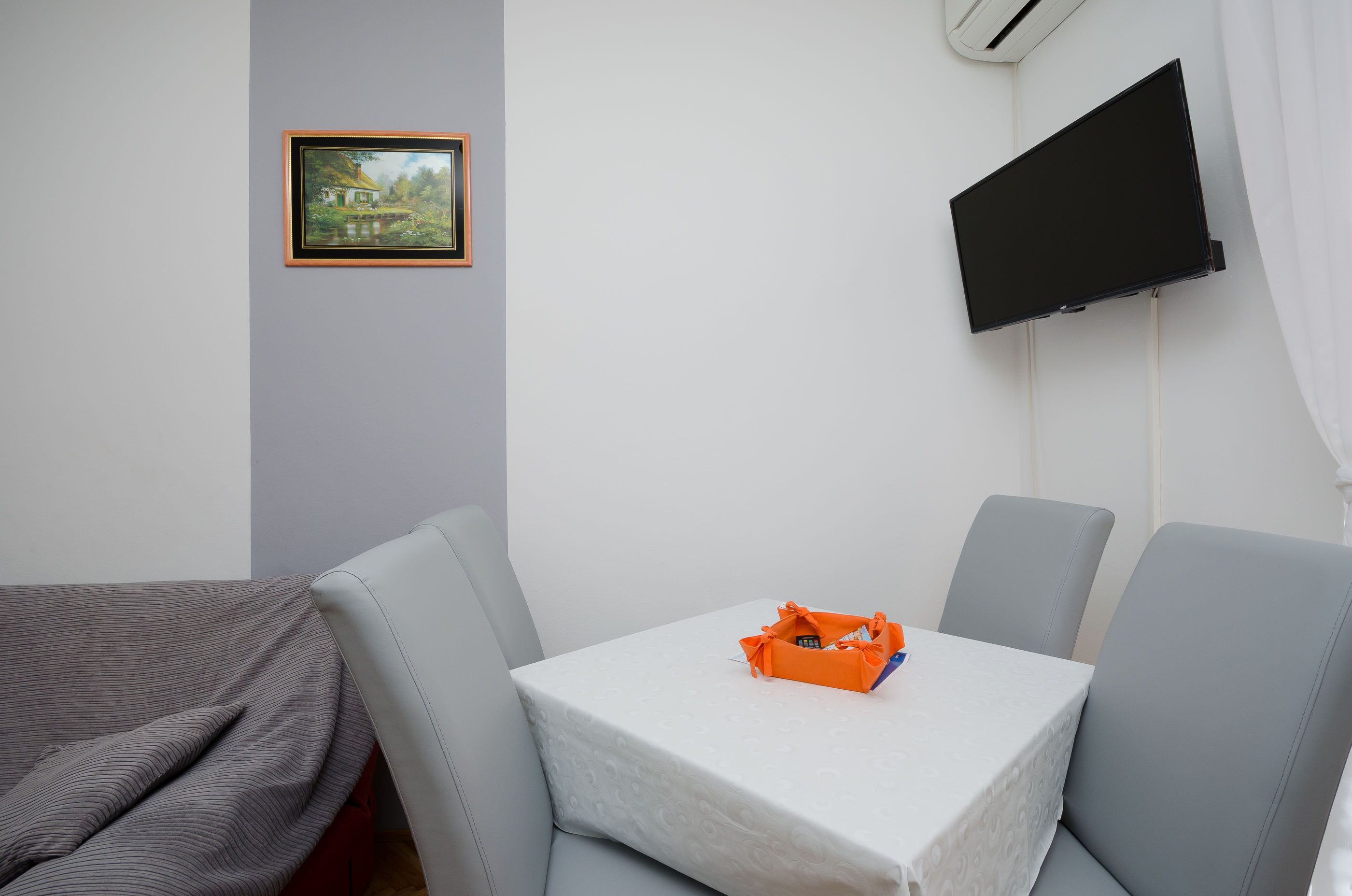 A hotel room with a bed, a small table with an orange basket, and a TV mounted on the wall.