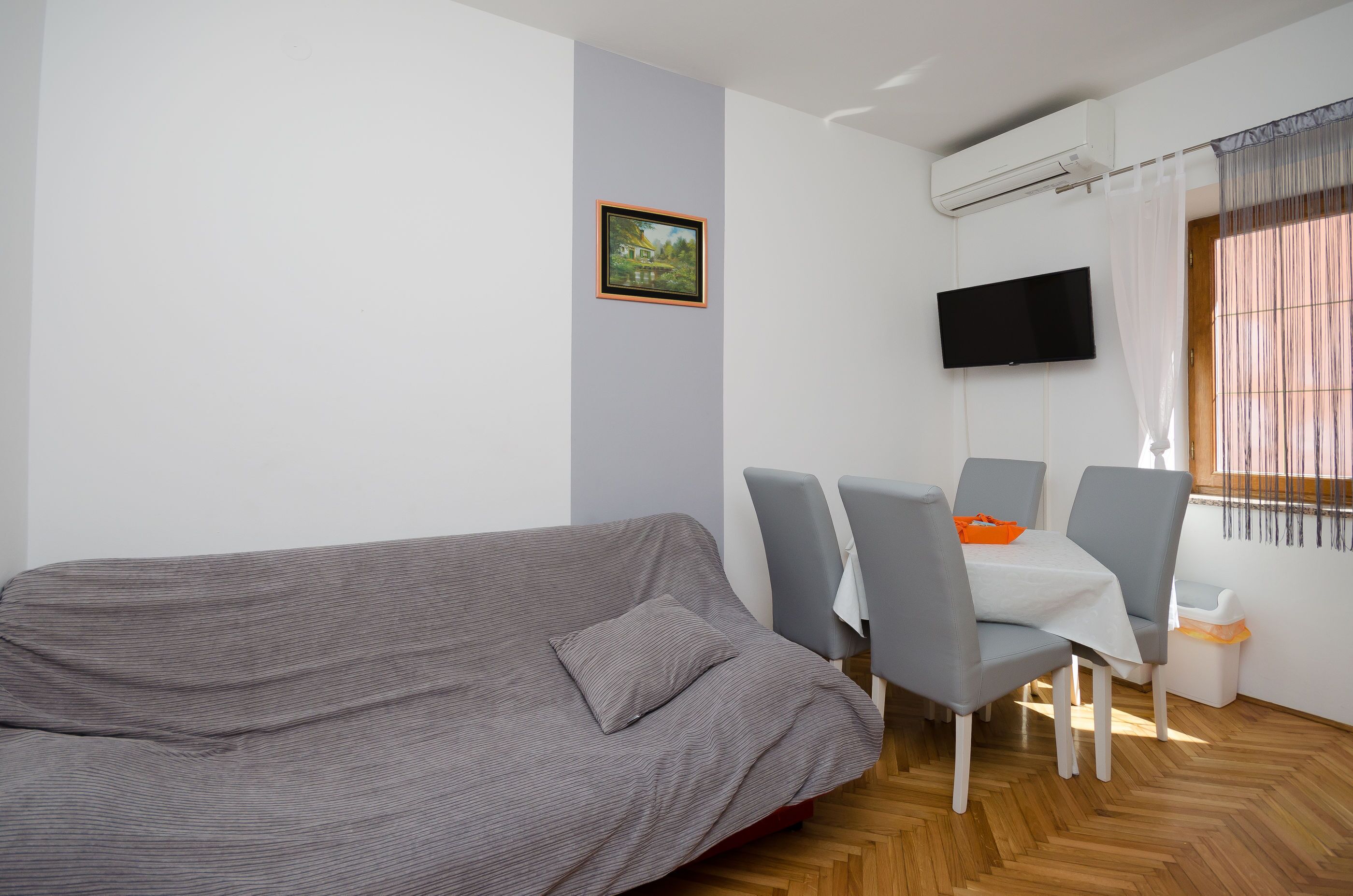 A modern living room with a grey sofa, a small table with chairs, a mounted TV, and a window with curtains.