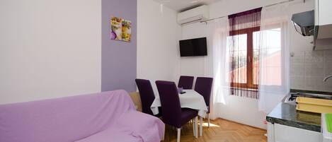 Une pièce de taille modeste, avec un canapé violet, une table à manger entourée de chaises violettes, un téléviseur fixé au mur et un coin cuisine équipé d’une cuisinière.