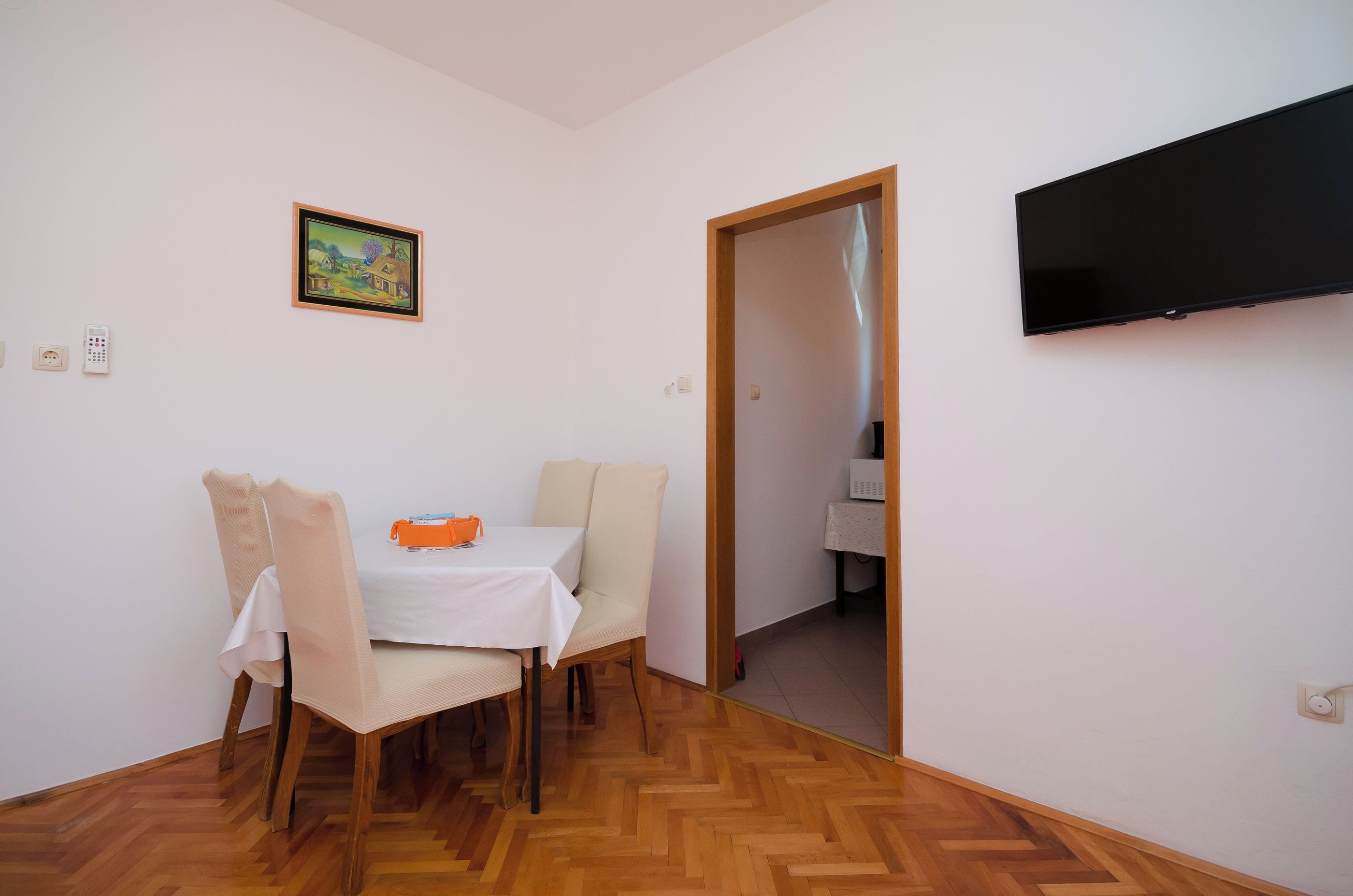 A dining area with a table set for two, a television mounted on the wall, and a door leading to another room.