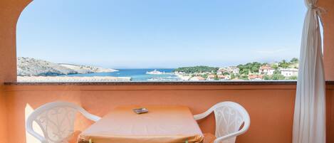 A balcony with a table and chairs, overlooking a coastal view with buildings and the sea.