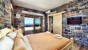 Un dormitorio con pared de piedra, una cama con ropa de cama color beige, un televisor montado en la pared y vistas al exterior.