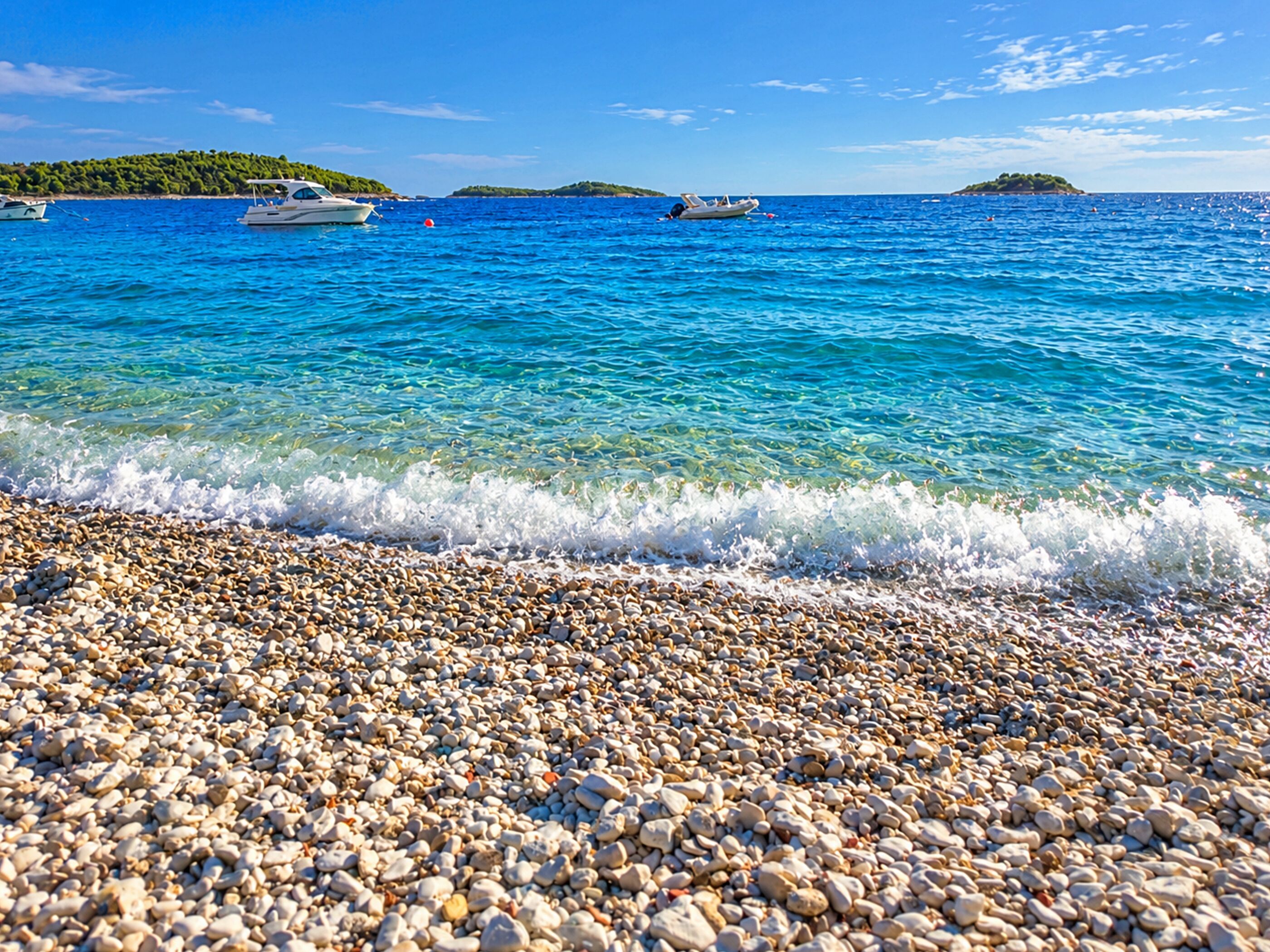 Une plage de galets bordée d’une eau d’un bleu limpide, avec des bateaux et des îles qui se dessinent à l’horizon.