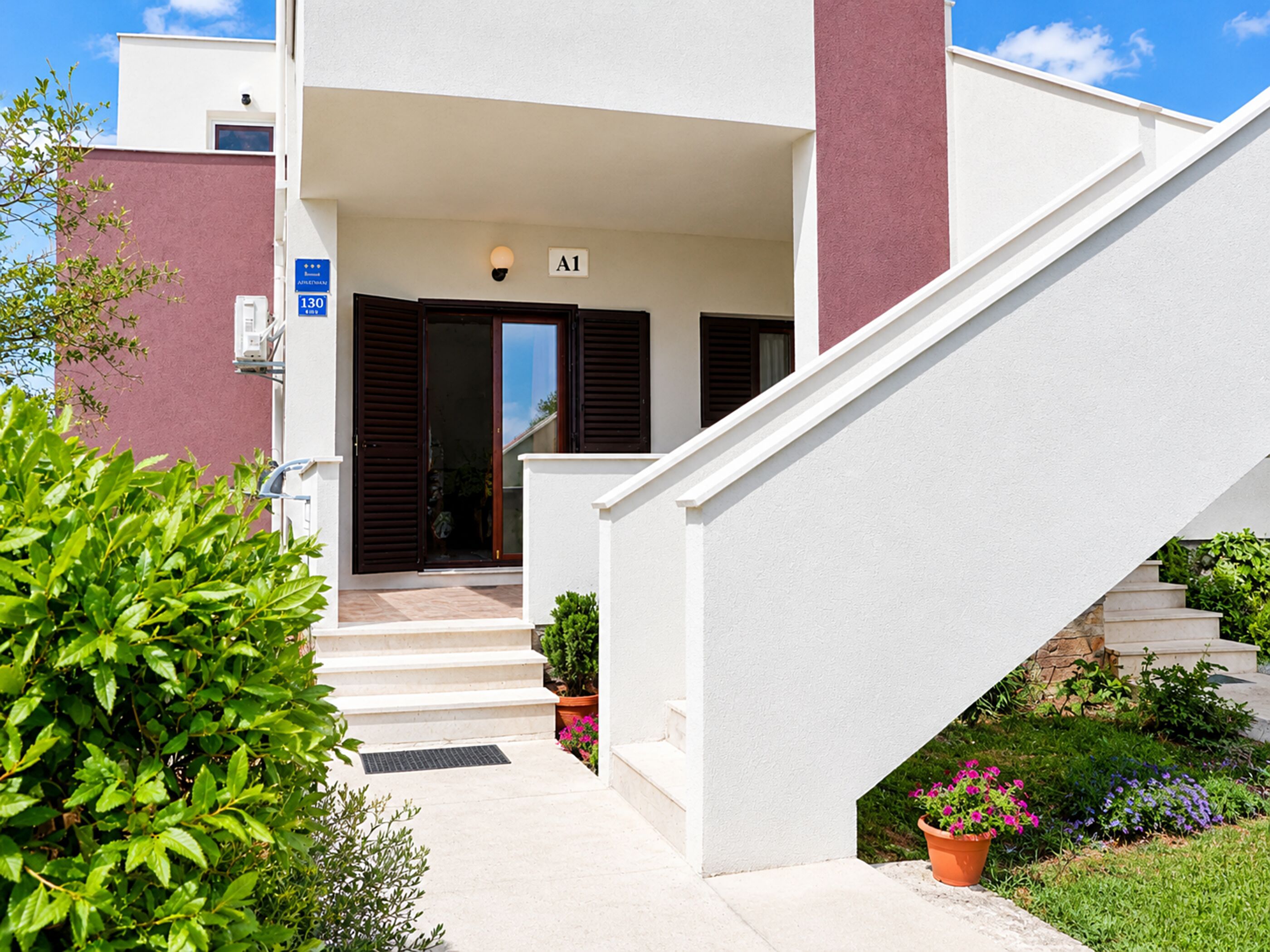 A modern building with a white exterior, a staircase leading to a door with a sign 'A1', and a small garden area with potted plants.